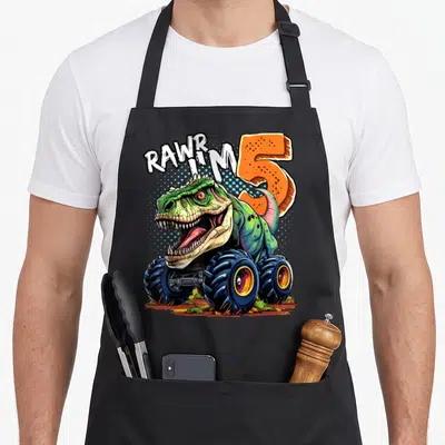 Rex 5th truck apron - Green dinosaur monster truck with white RAWR I'M text and orange number 5 — shown on a black cotton canvas apron against a white background