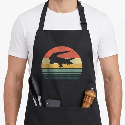 Alligator family aprons - Grey alligator silhouette over retro red orange yellow green blue grey striped sun — shown on a black cotton canvas apron against a white background