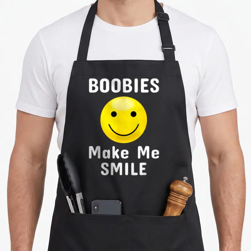 Booby mardi gras aprons - Yellow smiley face between white BOOBIES and Make Me SMILE text — shown on a black cotton canvas apron against a white background