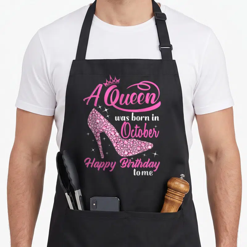 Retro october birthday apron - Pink high heel shoe with crown and A Queen was born in October text — shown on a black cotton canvas apron against a white background