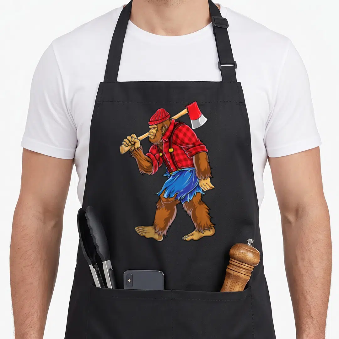 Bigfoot woodworking apron - Cartoon sasquatch in red plaid shirt and blue skirt carrying an axe over shoulder — shown on a black cotton canvas apron against a white background