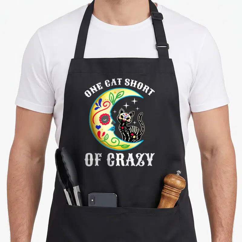 Sugar black cat aprons - Yellow moon profile facing black skeleton cat with white text ONE CAT SHORT OF CRAZY — shown on a black cotton canvas apron against a white background