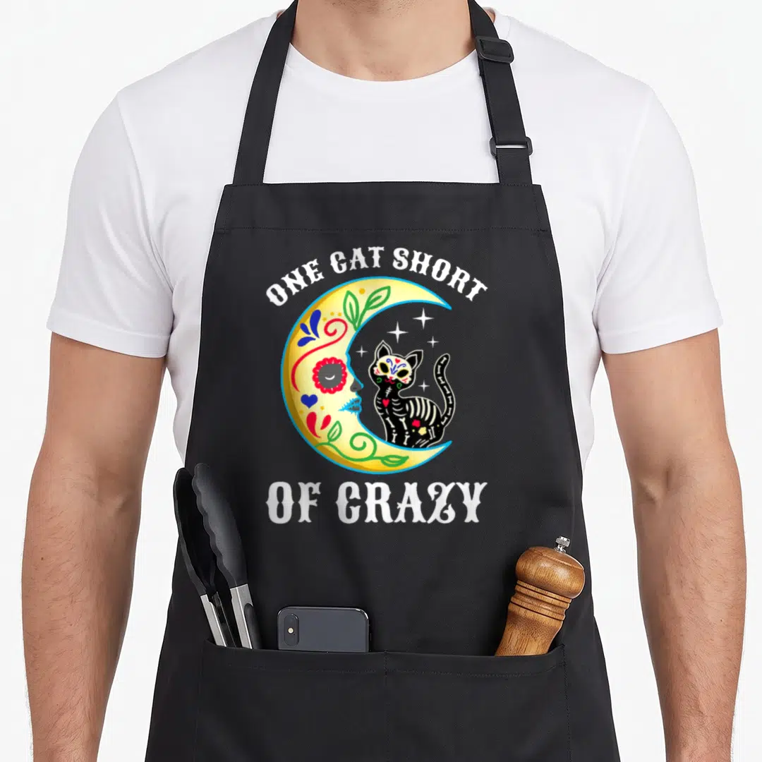 Sugar black cat aprons - Yellow moon profile facing black skeleton cat with white text ONE CAT SHORT OF CRAZY — shown on a black cotton canvas apron against a white background