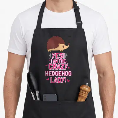 Outfit crazy hedgehog lady apron - Cartoon brown hedgehog above pink text YES I AM THE CRAZY HEDGEHOG LADY with hearts — shown on a black cotton canvas apron against a white background