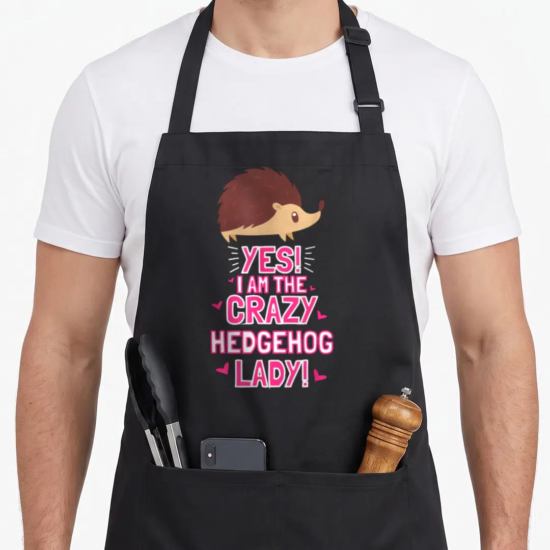 Outfit crazy hedgehog lady apron - Cartoon brown hedgehog above pink text YES I AM THE CRAZY HEDGEHOG LADY with hearts — shown on a black cotton canvas apron against a white background