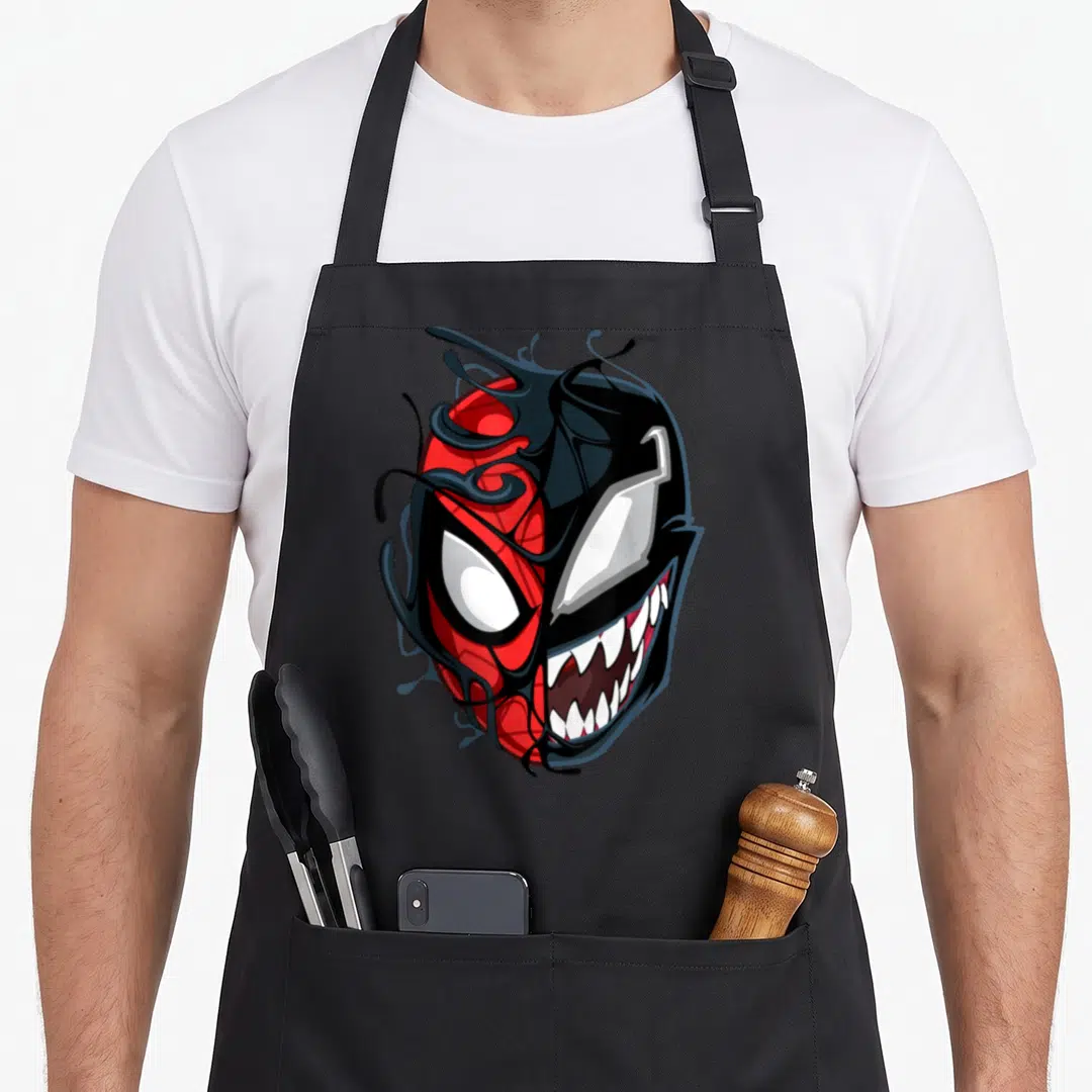 Maximum face apron - Split mask illustration in red, black, and white showing two distinct faces divided vertically — shown on a black cotton canvas apron against a white background