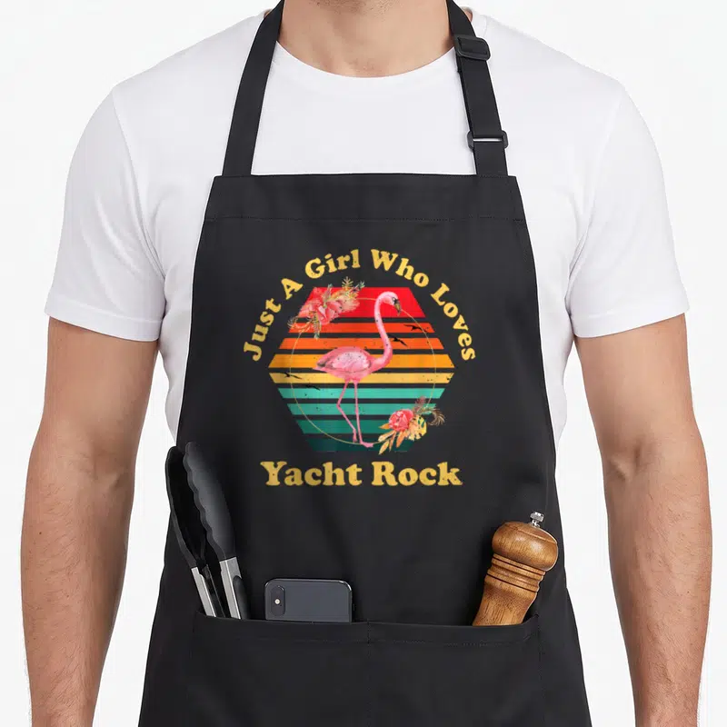 Yacht flamingo apron - Pink flamingo stands before striped sunset with Just A Girl Who Loves Yacht Rock text — shown on a black cotton canvas apron against a white background