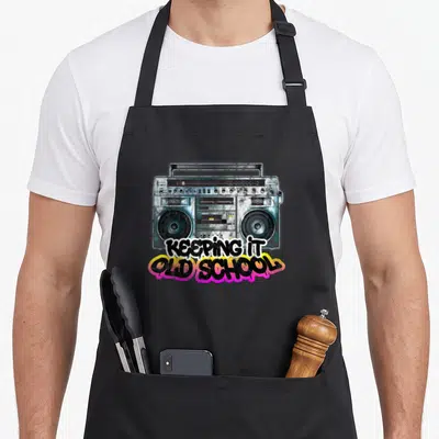 Keeping 80s apron - Realistic grey boombox with KEEPING IT OLD SCHOOL graffiti text in pink, yellow, and white — shown on a black cotton canvas apron against a white background