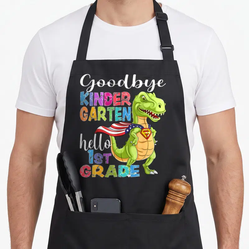 1st hello apron - Green T-Rex superhero with cape beside colorful Goodbye Kindergarten Hello 1st Grade text — shown on a black cotton canvas apron against a white background