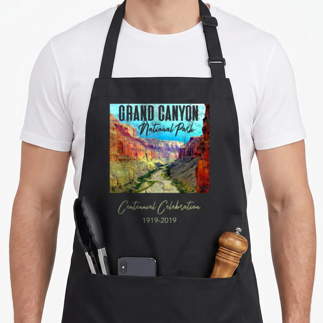 Grand hawthorne apron - Grand Canyon photo with red orange beige rocks and Centennial Celebration 1919-2019 text overlay — shown on a black cotton canvas apron against a white background
