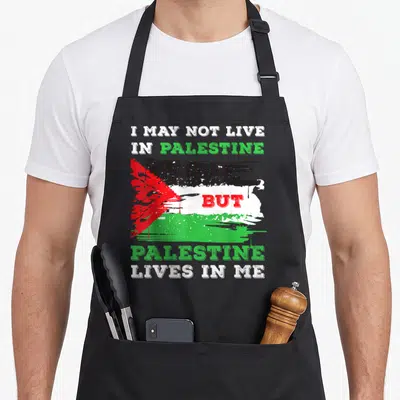 Flag live palestinian apron - Distressed Palestinian flag with text I MAY NOT LIVE IN PALESTINE BUT PALESTINE LIVES — shown on a black cotton canvas apron against a white background