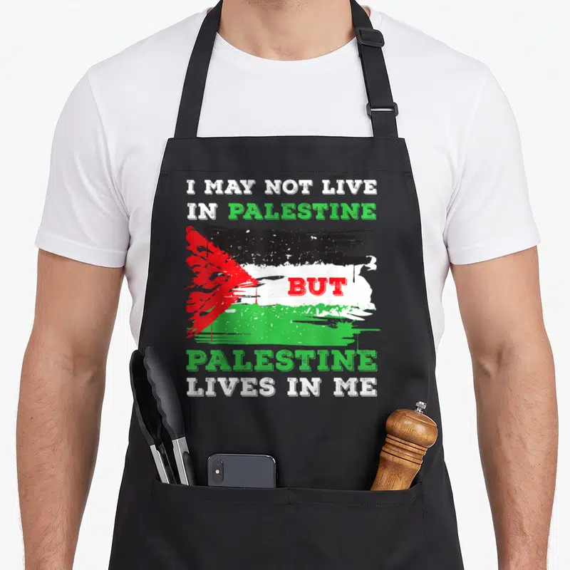 Flag live palestinian apron - Distressed Palestinian flag with text I MAY NOT LIVE IN PALESTINE BUT PALESTINE LIVES — shown on a black cotton canvas apron against a white background