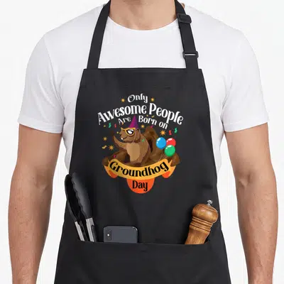 Funny groundhog birthday apron - Cartoon groundhog with party hat and balloons, text Only Awesome People Born — shown on a black cotton canvas apron against a white background