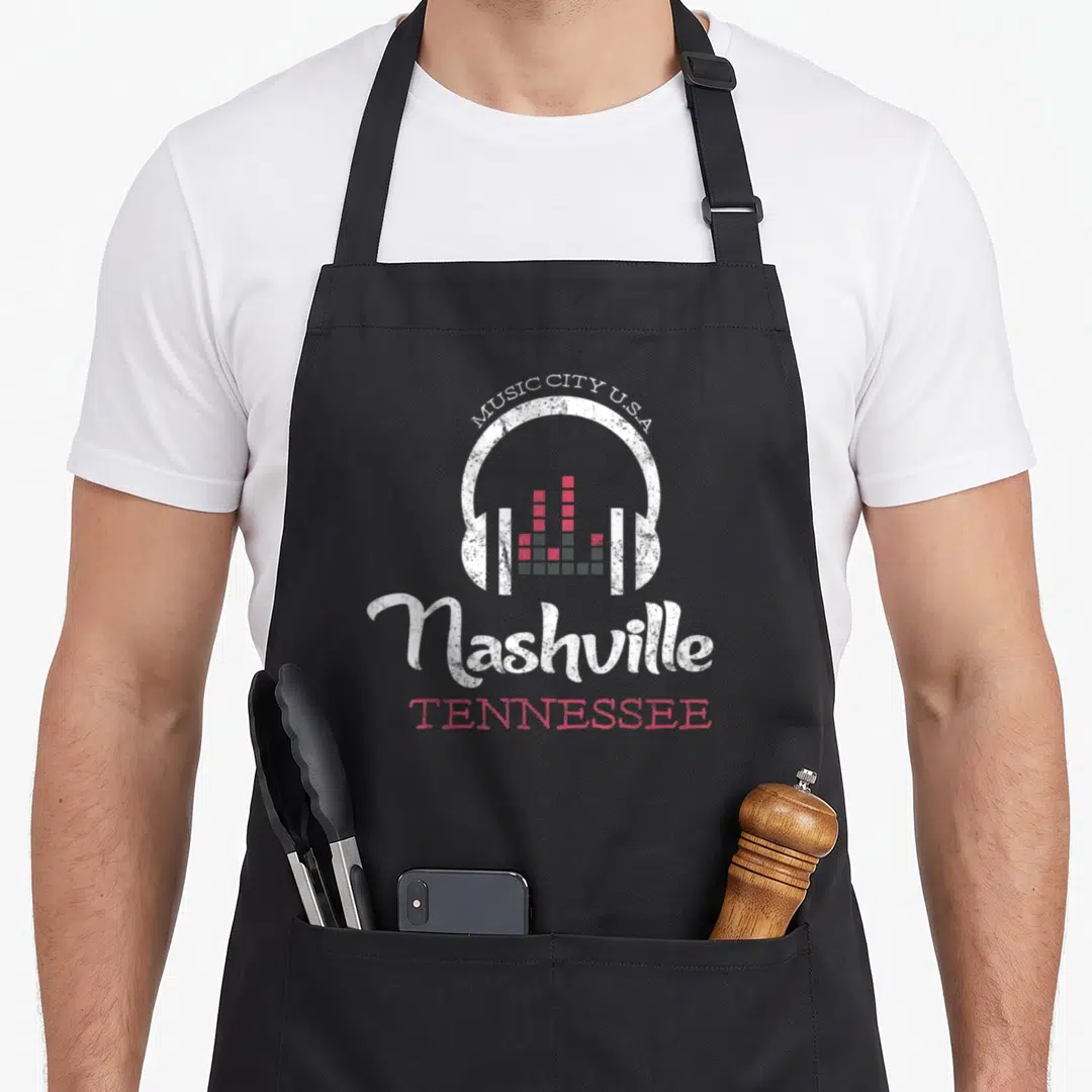 Nashville music country apron - Distressed white headphones with red and grey equalizer bars and Music City USA text — shown on a black cotton canvas apron against a white background