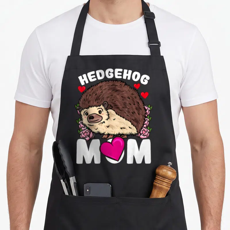Funny hedgehog apron - Cartoon brown hedgehog surrounded by pink roses and green leaves with white HEDGEHOG MOM text — shown on a black cotton canvas apron against a white background