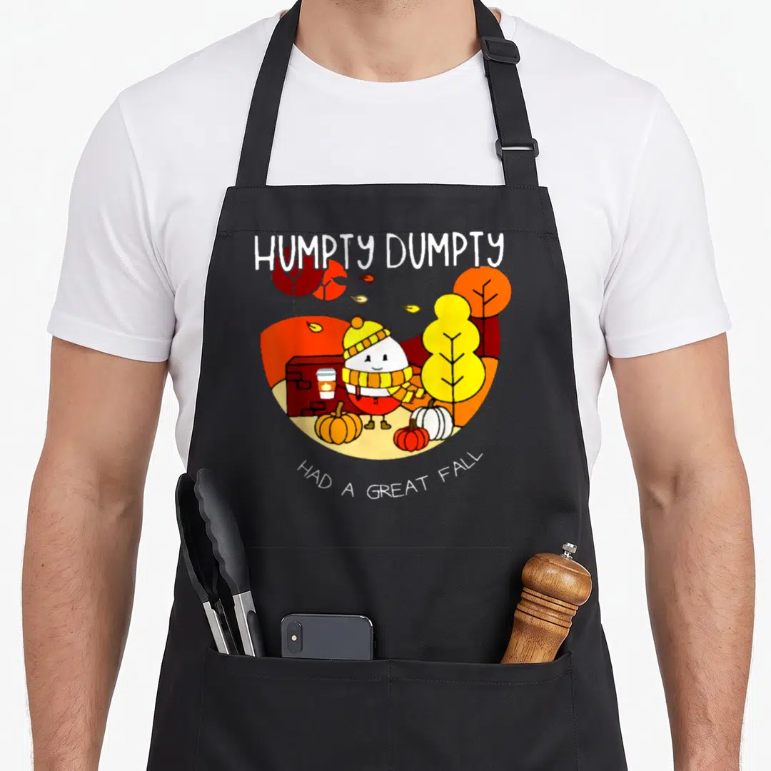 Retro humpty cute fall aprons - Cartoon Humpty Dumpty in scarf standing among orange and white pumpkins near a brick — shown on a black cotton canvas apron against a white background