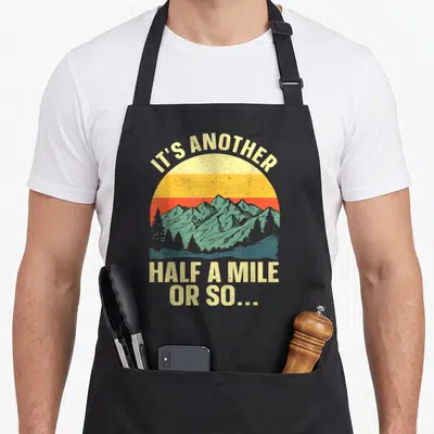 Backpacking mountain camping apron - Retro sun graphic with teal mountains and trees and beige text reading It's — shown on a black cotton canvas apron against a white background