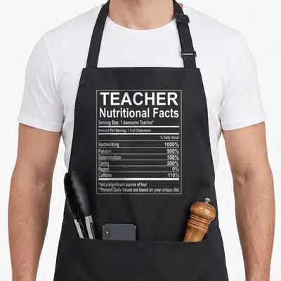 Nutrition teacher apron - White nutritional facts label graphic listing teacher traits like hardworking 1000% — shown on a black cotton canvas apron against a white background