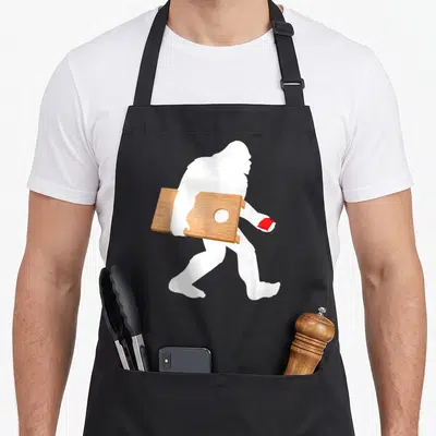Lawn game aprons - White bigfoot silhouette carrying brown cornhole board and red bean bag — shown on a black cotton canvas apron against a white background