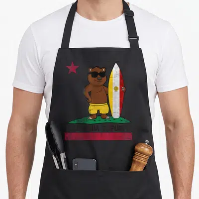 San bear apron - Cartoon brown bear in yellow shorts holding a surfboard with CALIFORNIA REPUBLIC text and red star — shown on a black cotton canvas apron against a white background