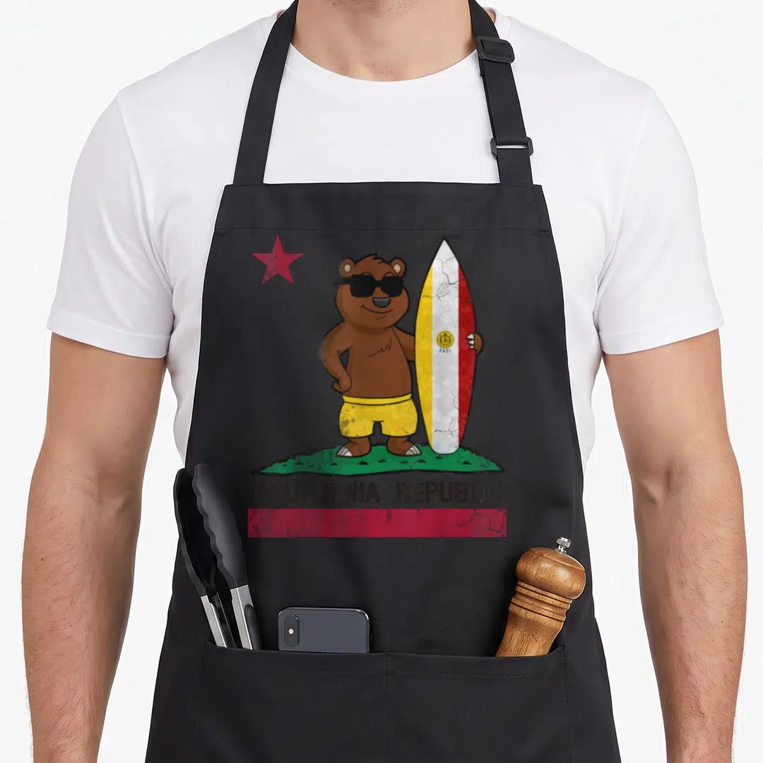 San bear apron - Cartoon brown bear in yellow shorts holding a surfboard with CALIFORNIA REPUBLIC text and red star — shown on a black cotton canvas apron against a white background