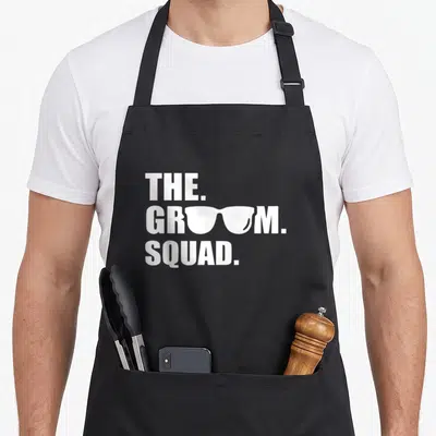 Squad groom apron - White stacked text THE GROOM SQUAD with sunglasses replacing vowels on center line — shown on a black cotton canvas apron against a white background