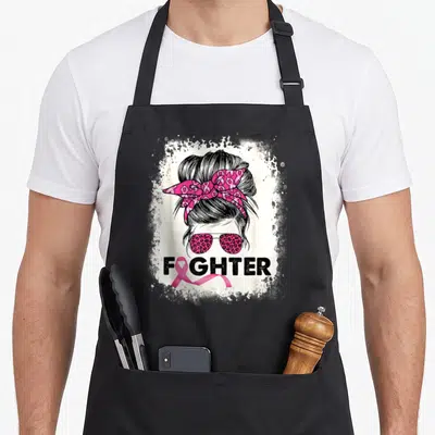 Bun breast cancer awareness aprons - Stylized messy bun with pink ribbon bandana, leopard sunglasses, and FIGHTER text — shown on a black cotton canvas apron against a white background