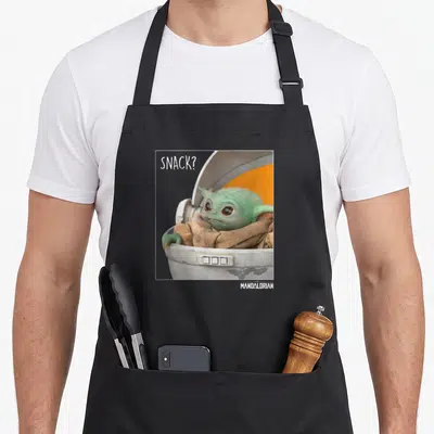 Funny snack time apron - Grogu in a pod with SNACK text and white frame border — shown on a black cotton canvas apron against a white background