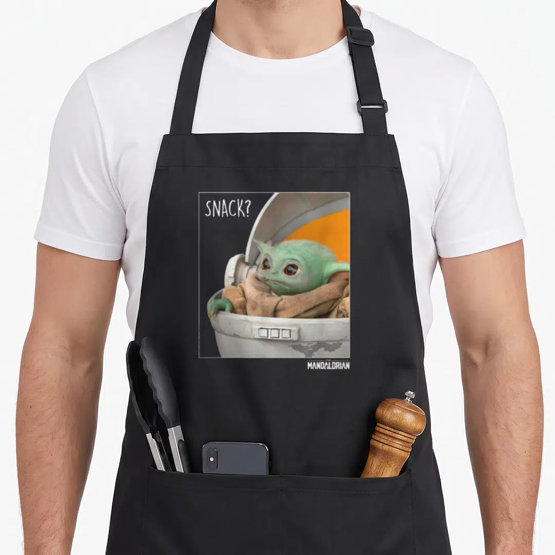 Funny snack time apron - Grogu in a pod with SNACK text and white frame border — shown on a black cotton canvas apron against a white background