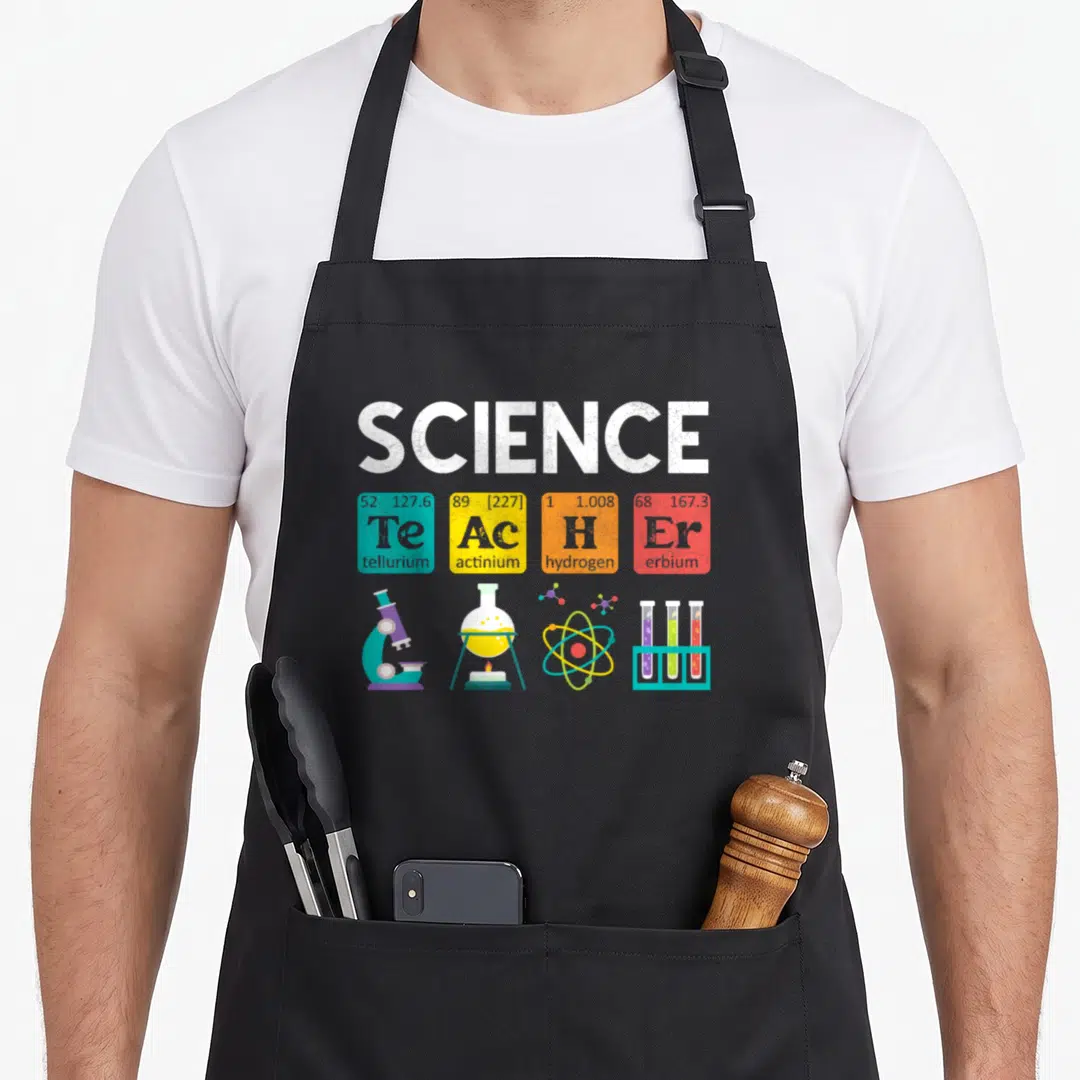 Biology chemistry apron - Periodic table tiles spelling teacher with microscope flask atom and test tubes in cartoon — shown on a black cotton canvas apron against a white background
