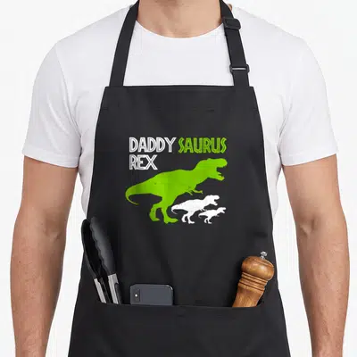 Rex father day apron - DADDY SAURUS REX text above three green and white T-Rex silhouettes facing right — shown on a black cotton canvas apron against a white background