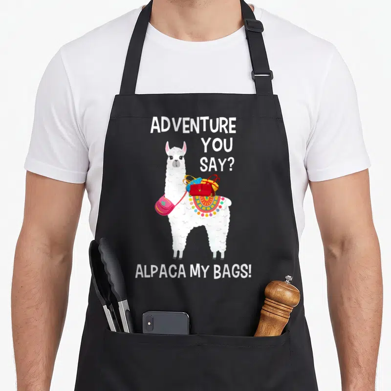 Bag alpaca apron - White alpaca wearing red blue and yellow bags with punny text — shown on a black cotton canvas apron against a white background