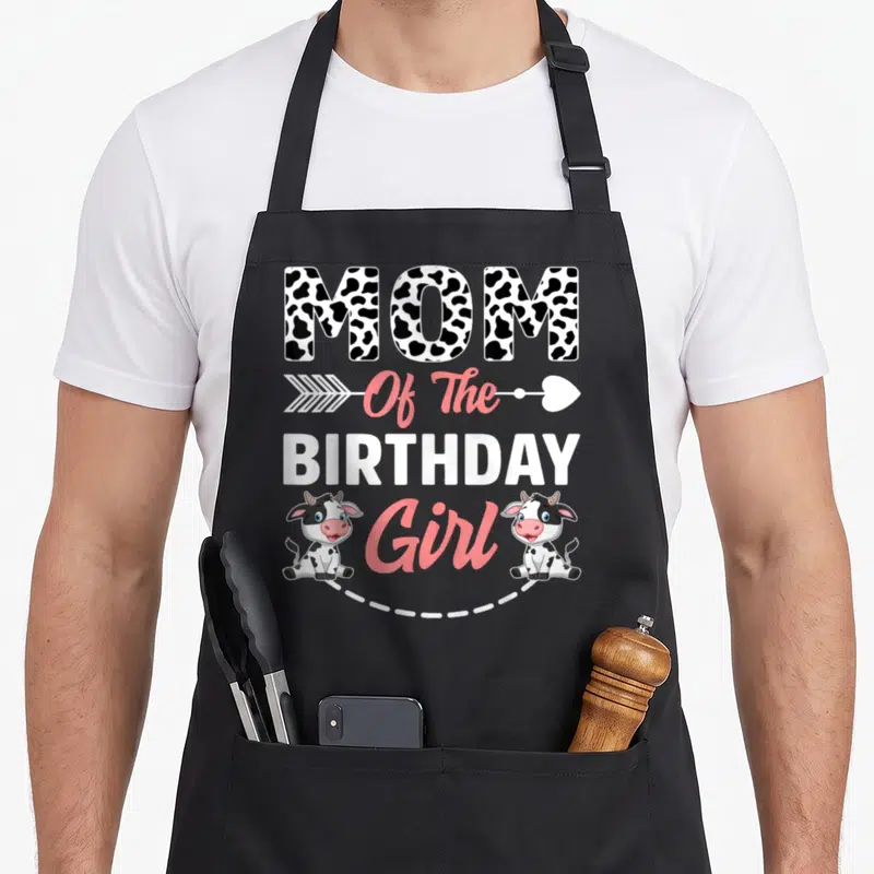 Animal cow print apron - MOM Of The BIRTHDAY Girl text with two cartoon cows and pink arrow — shown on a black cotton canvas apron against a white background