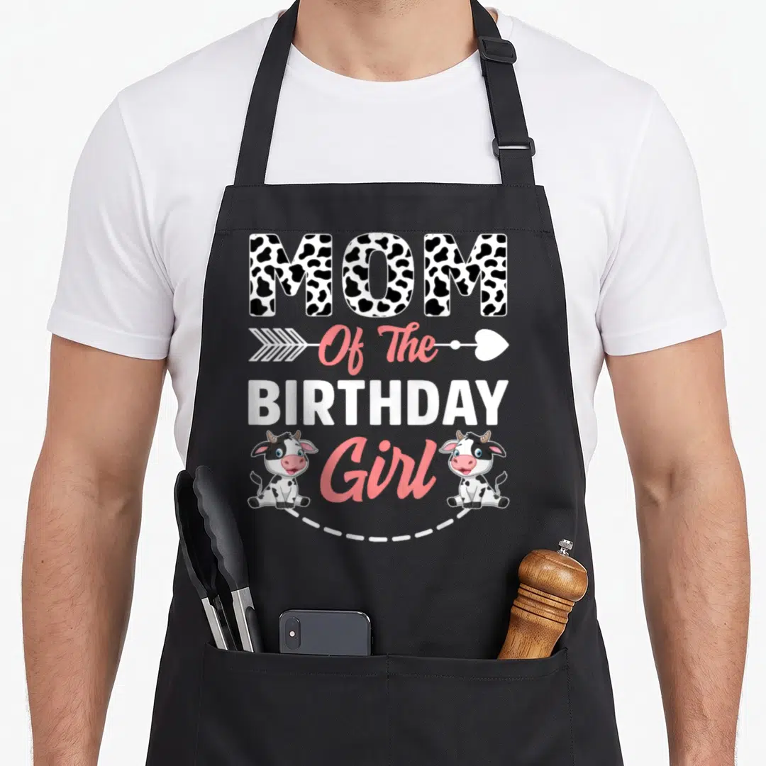 Animal cow print apron - MOM Of The BIRTHDAY Girl text with two cartoon cows and pink arrow — shown on a black cotton canvas apron against a white background