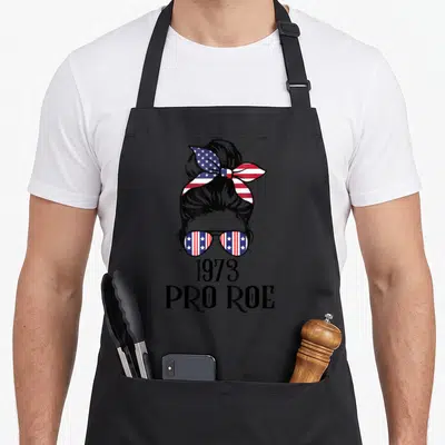 Pro messy aprons - 1973 PRO ROE text below cartoon woman's bun and stars stripes sunglasses — shown on a black cotton canvas apron against a white background