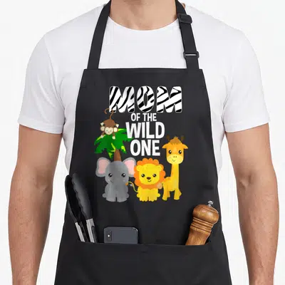 Safari birthday apron - MOM OF THE WILD ONE text with cartoon monkey, elephant, lion, giraffe and palm tree — shown on a black cotton canvas apron against a white background