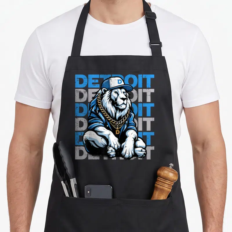 Youth lion apron - Cartoon lion in cap and gold chain before five stacked Detroit text lines in blue and grey — shown on a black cotton canvas apron against a white background
