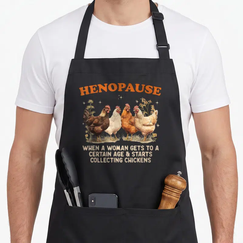 Henopause chicken apron - Four chickens standing in a row under arched orange text reading HENOPAUSE with three lines — shown on a black cotton canvas apron against a white background