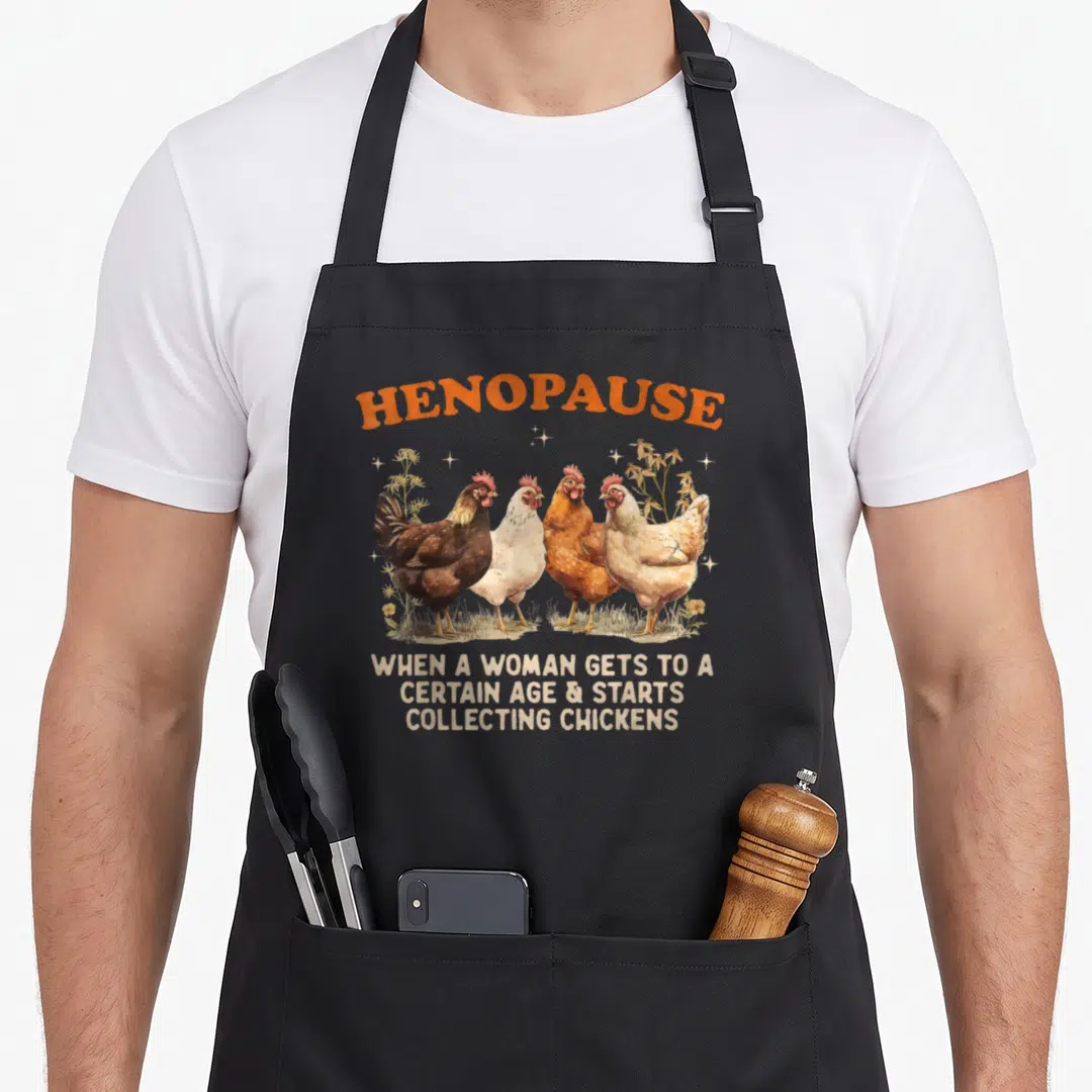 Henopause chicken apron - Four chickens standing in a row under arched orange text reading HENOPAUSE with three lines — shown on a black cotton canvas apron against a white background