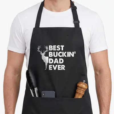 Buckin hockey dad apron - White deer profile next to stacked text BEST BUCKIN DAD EVER in distressed style — shown on a black cotton canvas apron against a white background