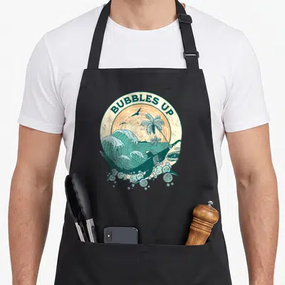 Shark bubble apron - Retro circular design with BUBBLES UP text, leaping shark, palm trees, and teal wave — shown on a black cotton canvas apron against a white background