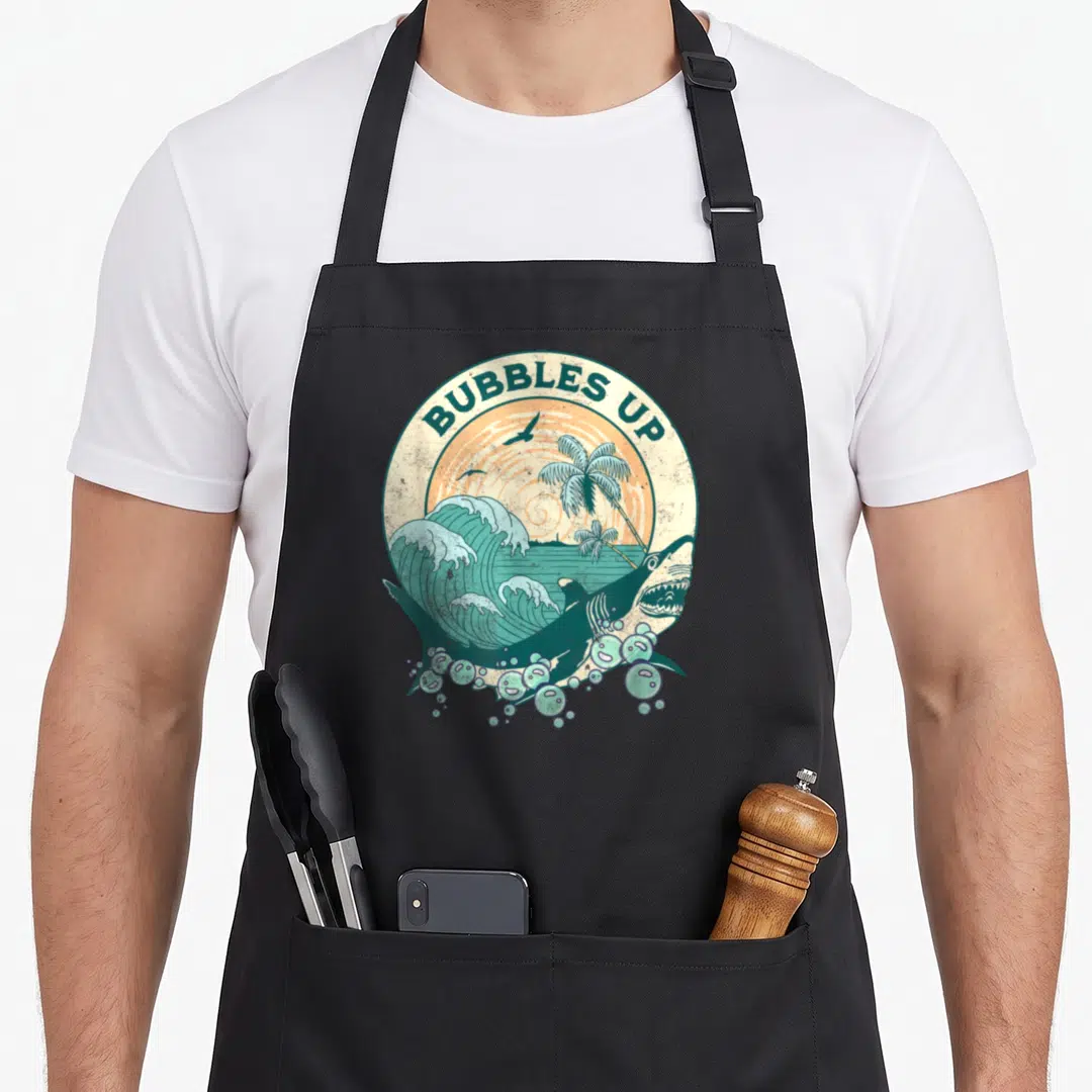 Shark bubble apron - Retro circular design with BUBBLES UP text, leaping shark, palm trees, and teal wave — shown on a black cotton canvas apron against a white background