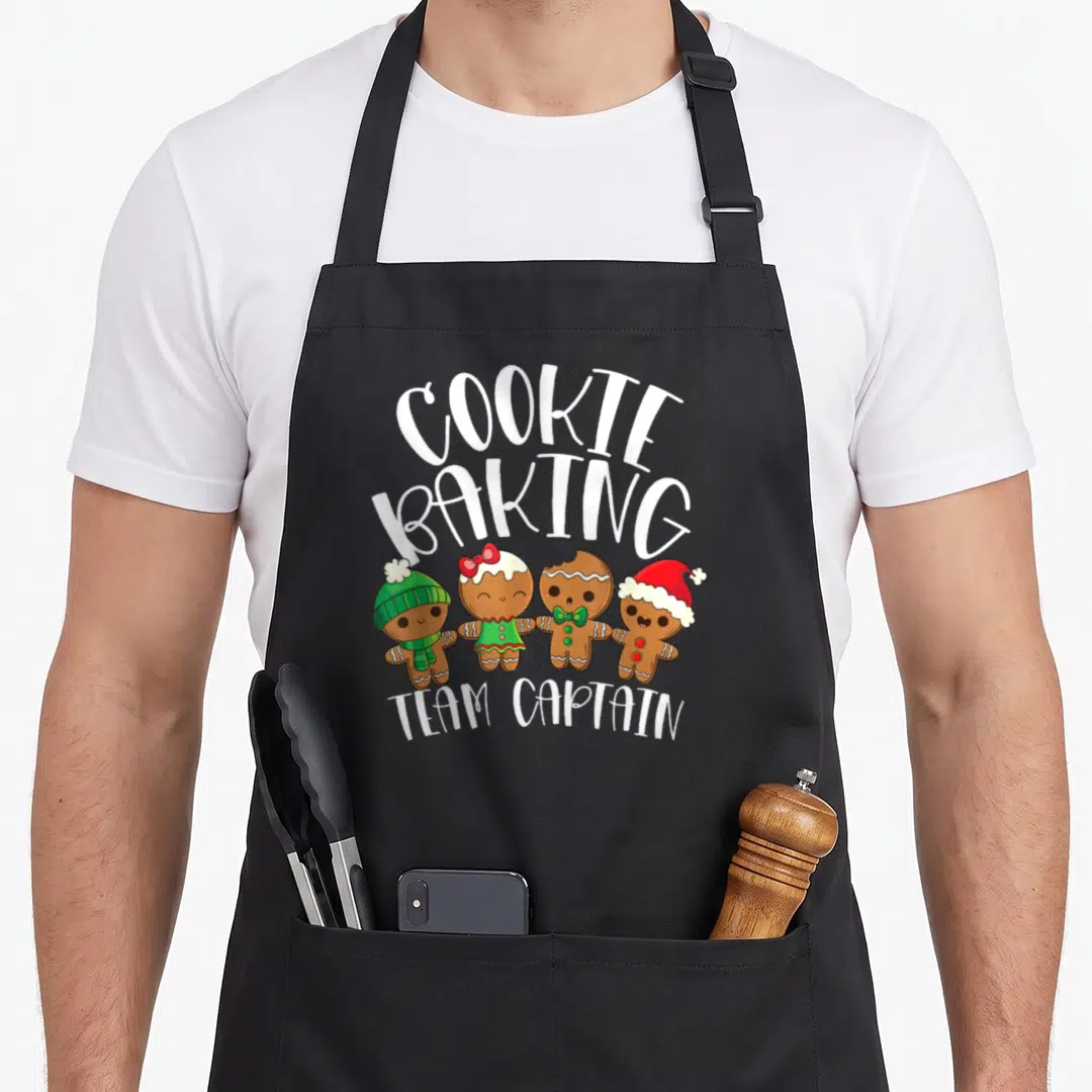 Captain baker apron - Four gingerbread men holding hands under arched Cookie Baking Team Captain text — shown on a black cotton canvas apron against a white background