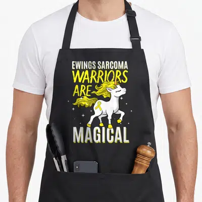 Warrior sarcoma awareness apron - Cartoon white unicorn yellow mane Ewings Sarcoma text yellow ribbon stars — shown on a black cotton canvas apron against a white background
