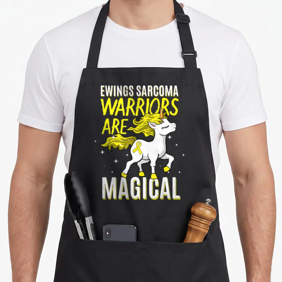 Warrior sarcoma awareness apron - Cartoon white unicorn yellow mane Ewings Sarcoma text yellow ribbon stars — shown on a black cotton canvas apron against a white background