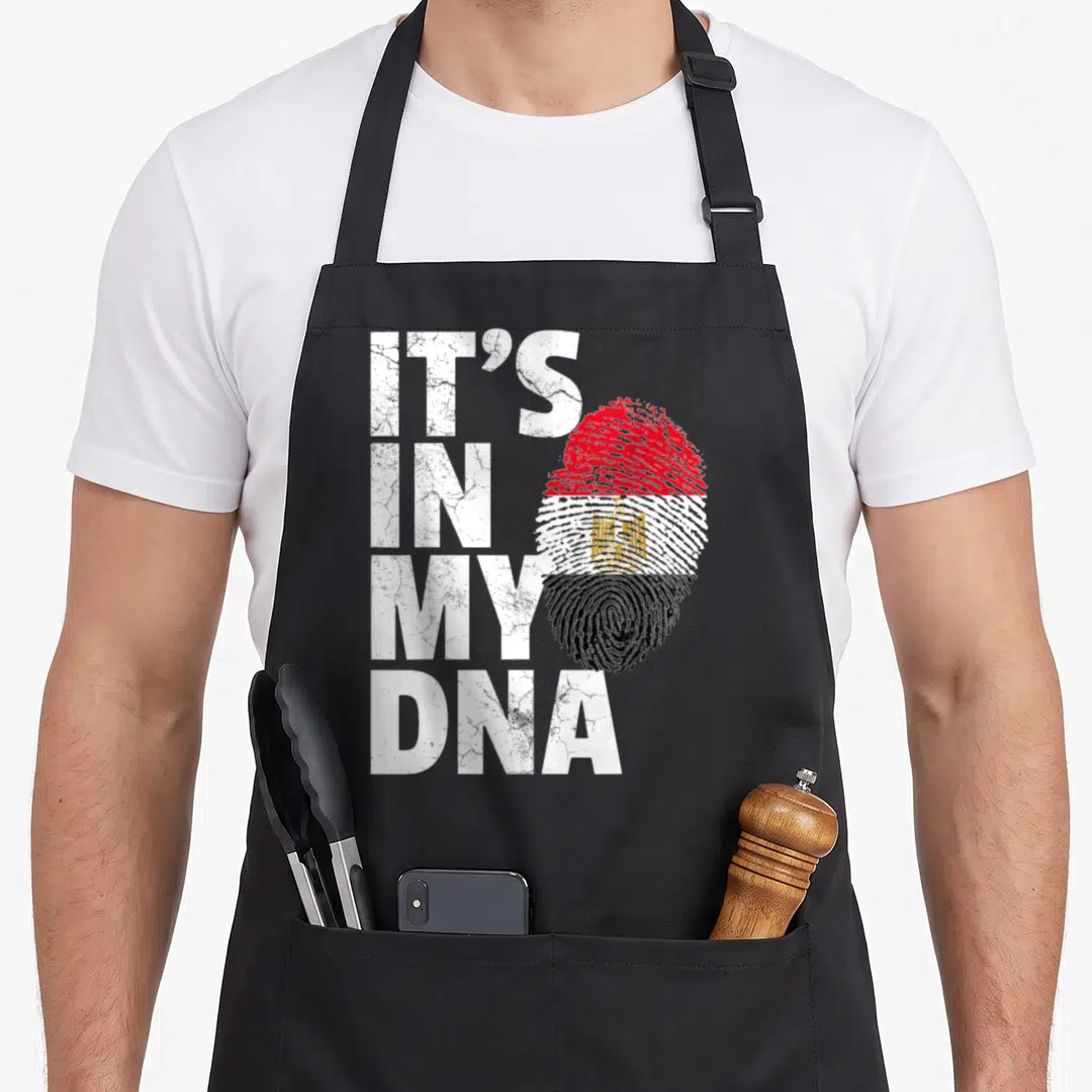 Dna egyptian apron - Distressed white stacked text next to a fingerprint shaped like the red white black and gold — shown on a black cotton canvas apron against a white background