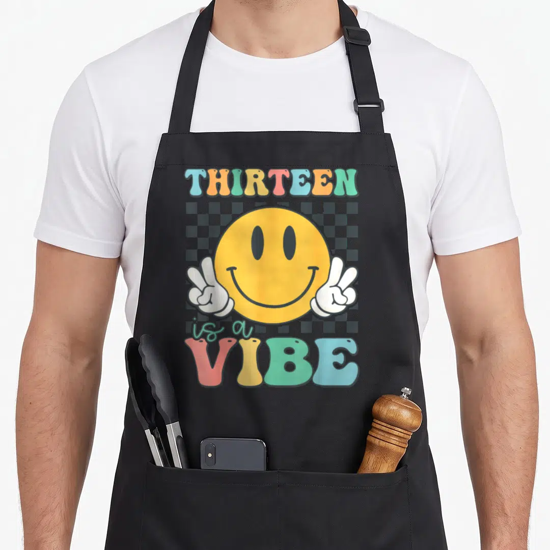 Thirteen custom face apron - Retro yellow smiley face with peace hands and checkerboard background under colorful text — shown on a black cotton canvas apron against a white background