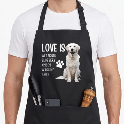Golden english aprons - White dog photo beside stacked text listing wet noses slobbery kisses and wagging tails — shown on a black cotton canvas apron against a white background