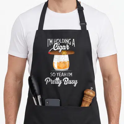 Whiskey smoking apron - Cigar balanced on whiskey glass with sarcastic text I'M HOLDING A CIGAR SO YEAH I'M PRETTY BUSY — shown on a black cotton canvas apron against a white background