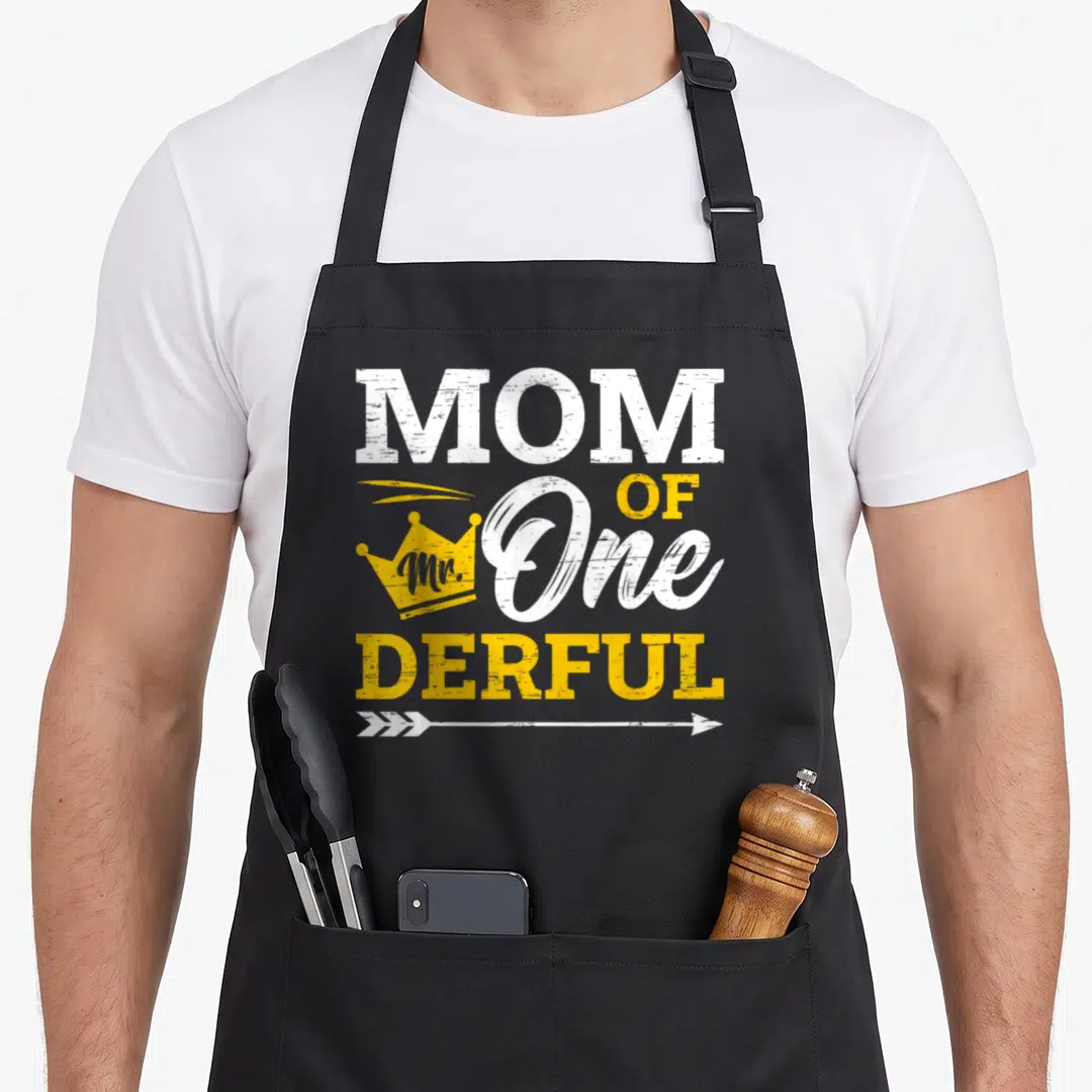 Natal birthday apron - MOM OF One Mr DERFUL text with yellow crown and white arrow on black background — shown on a black cotton canvas apron against a white background